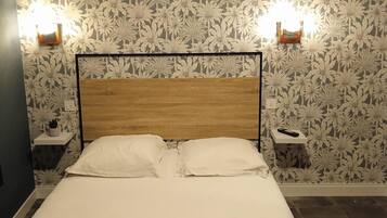 A neatly made bed with two pillows and two folded towels, a wooden headboard, and a floral wallpaper pattern.