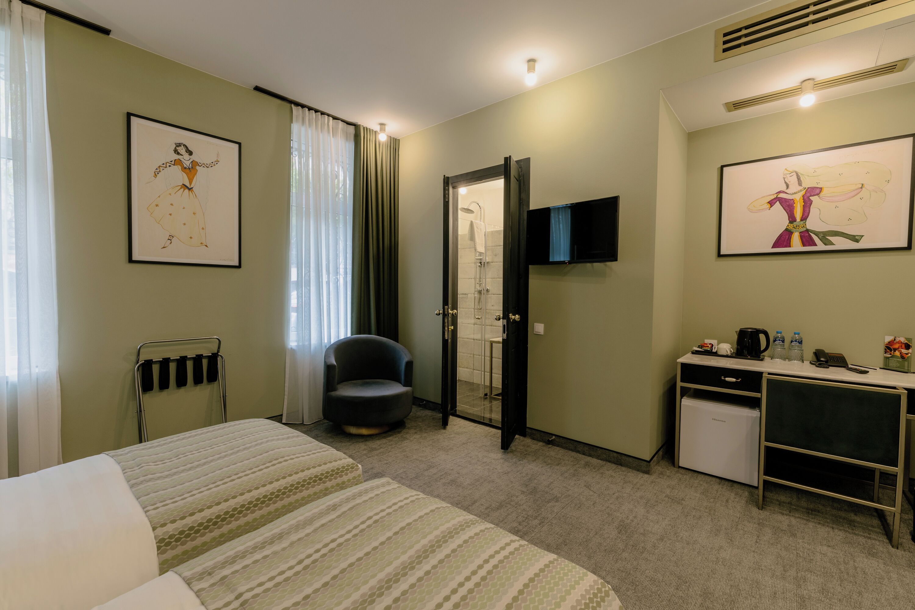 Habitación de hotel con cama, una silla, escritorio con computadora, ducha y dos cuadros en la pared.