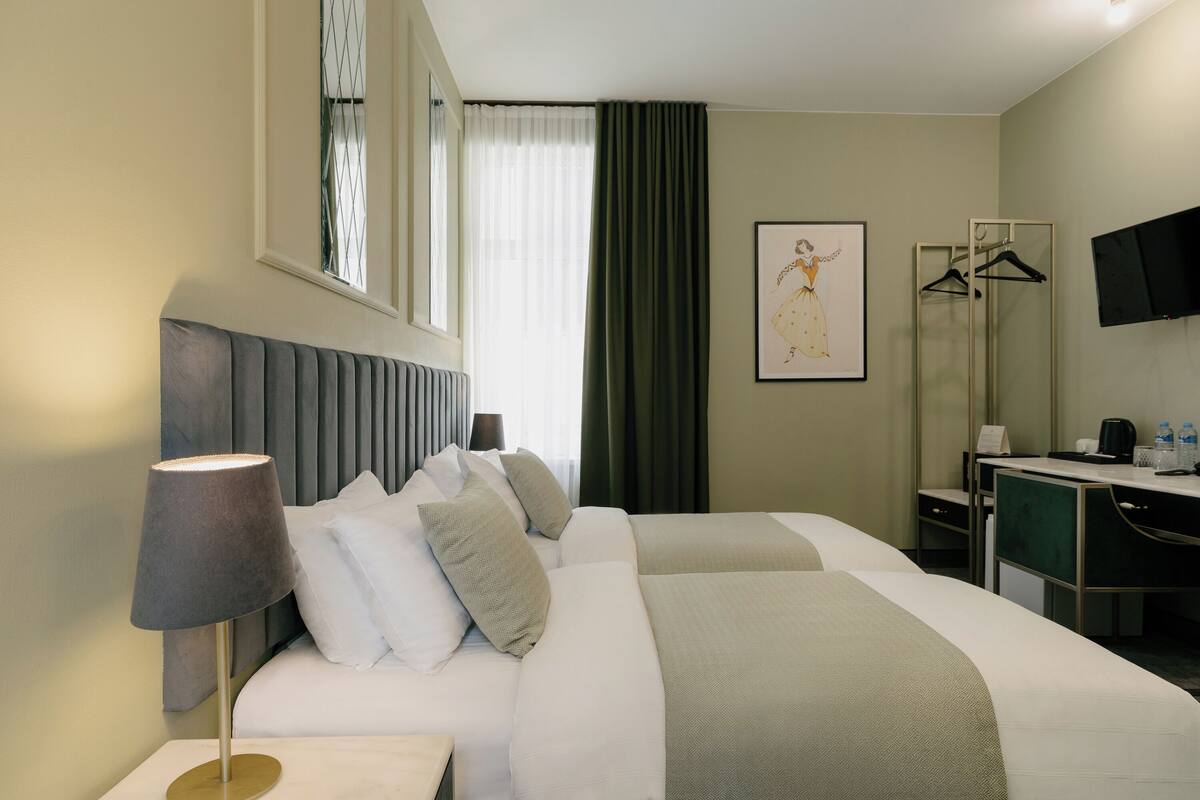 Una habitació d'hotel amb dos llits, escriptori, mirall, televisió i finestra amb cortines.