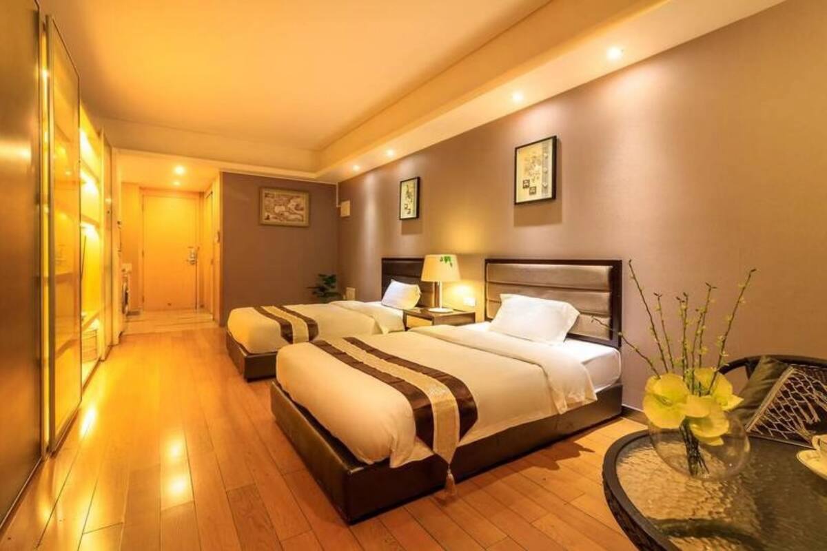 Ein Hotelzimmer mit zwei Betten, einem Nachttisch mit Lampe, einem kleinen runden Tisch mit Blumenvase und einem Flur, der zu einem weiteren Zimmer führt.