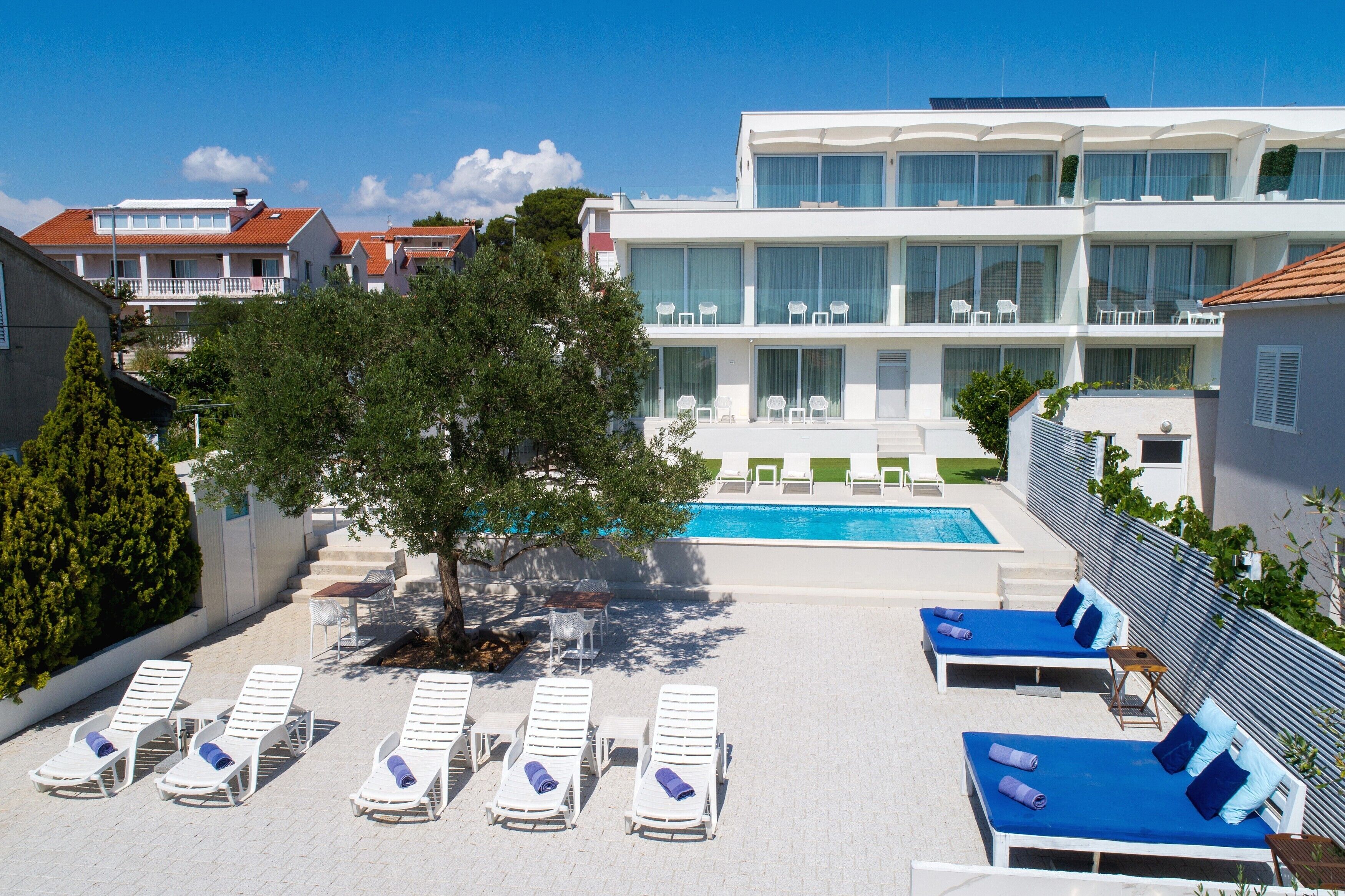 A rooftop pool area with sun loungers and a swimming pool, surrounded by modern buildings and a tree.