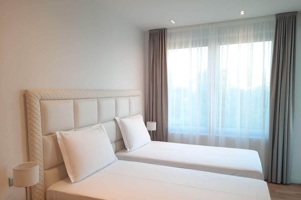 A hotel room with two beds, a nightstand, a lamp, and a large window with curtains.