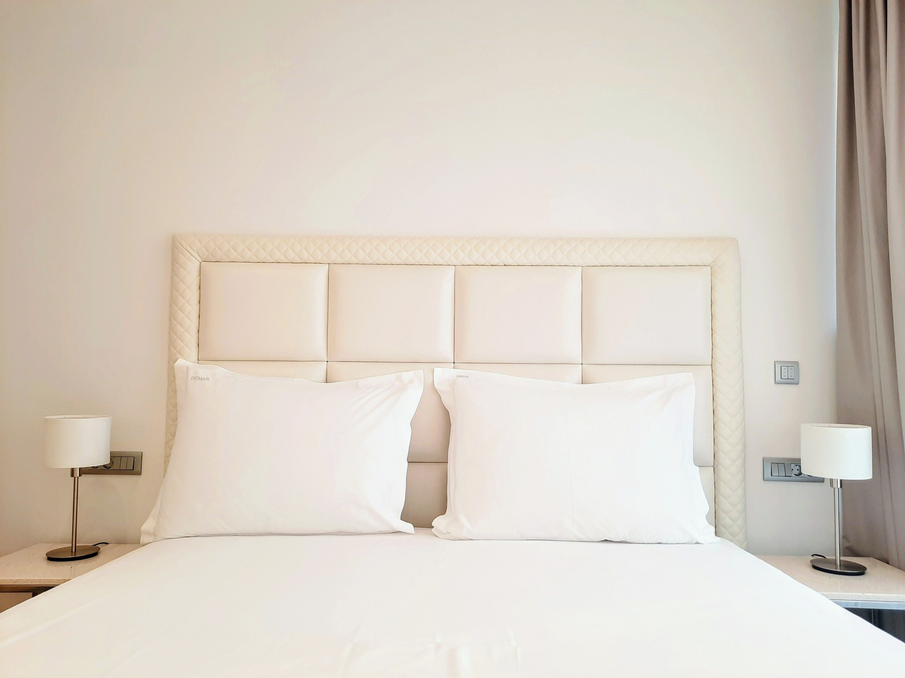 A neatly made bed with a quilted headboard, two bedside tables with lamps, and a power outlet on the wall.