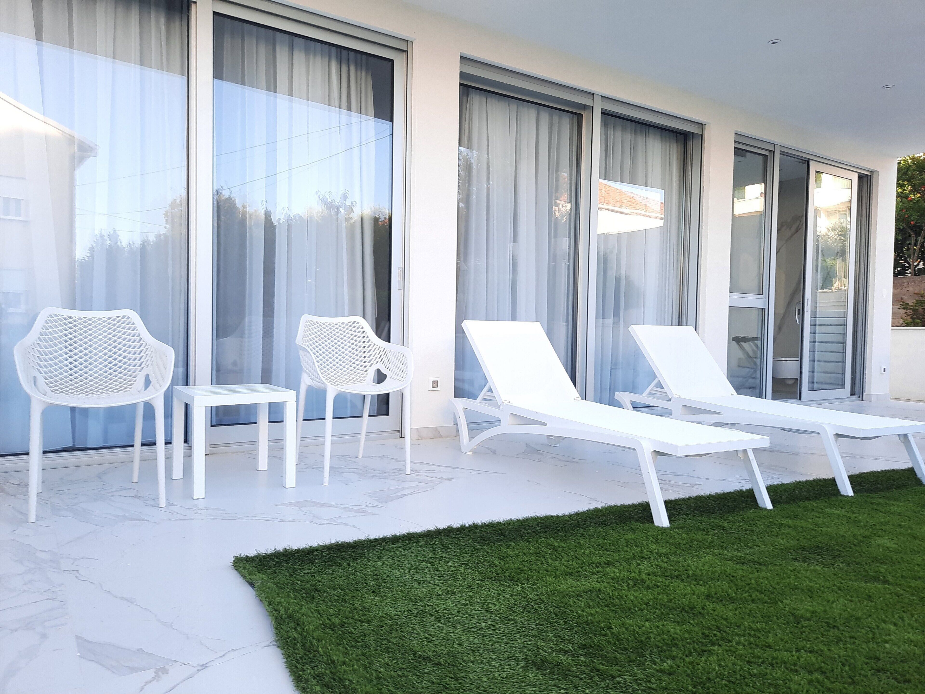A modern outdoor seating area with white chairs and lounge chairs, large glass doors, and a green artificial lawn.