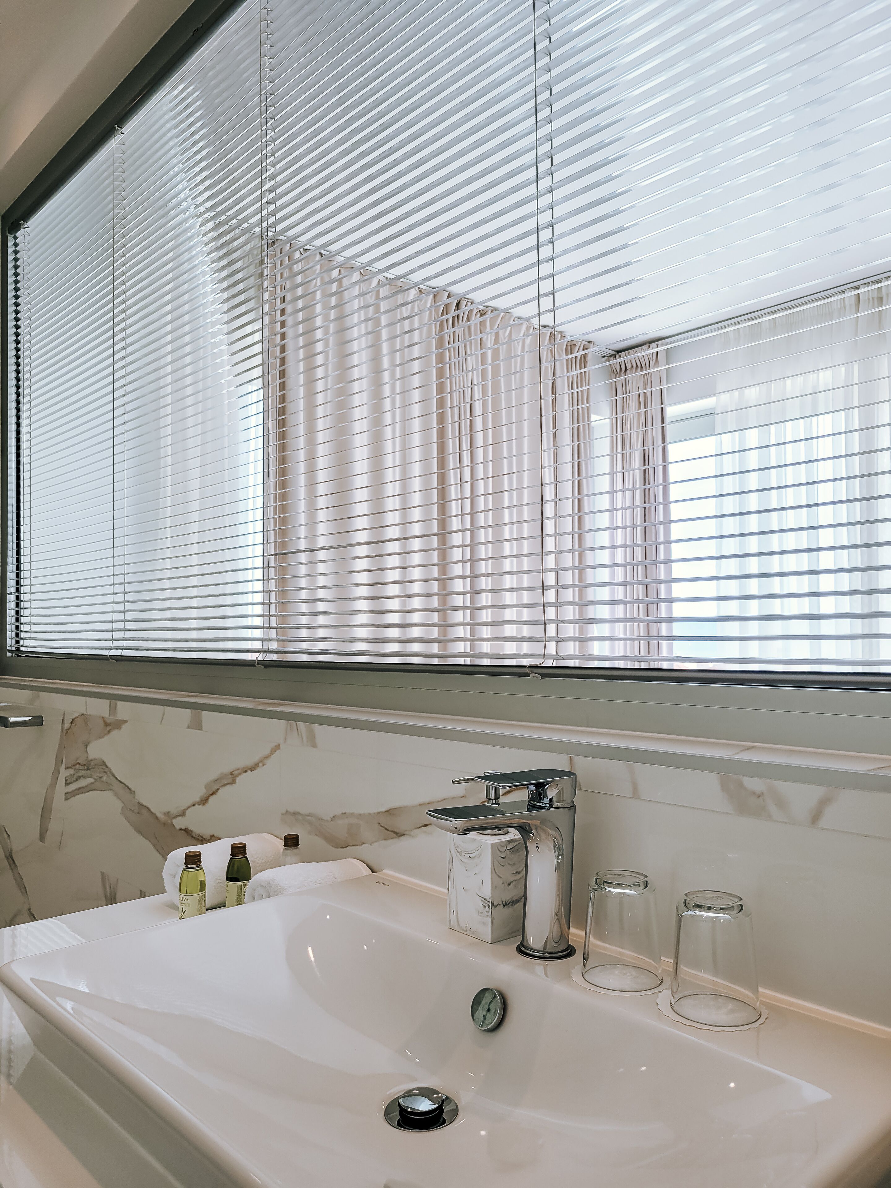 Un baño con un espejo grande, un lavamanos de mármol y una ventana con persianas.