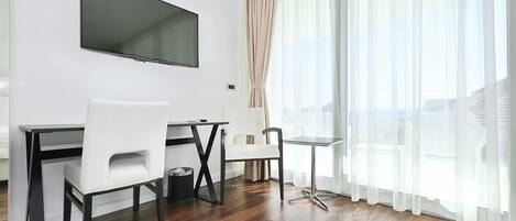 A modern hotel room with a flat-screen TV mounted on the wall, a wooden floor, two white chairs, a small round table, and large windows with sheer curtains.
