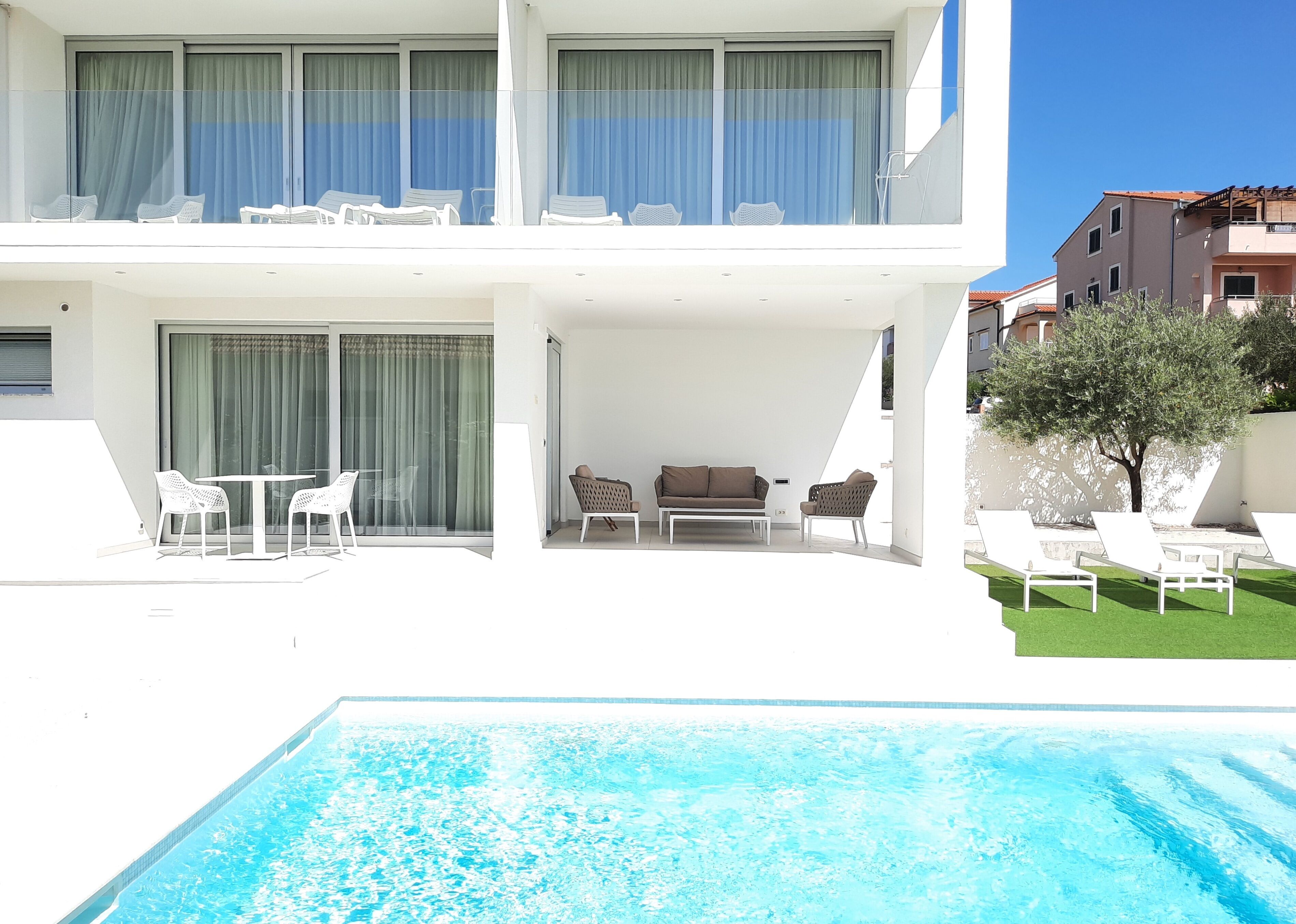 A modern two-story building with a pool, outdoor seating, and a tree.