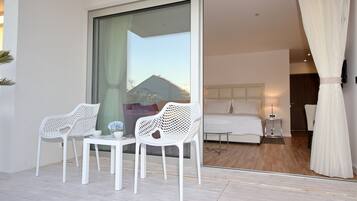 A modern hotel room with a large sliding glass door leading to a balcony. The room features a bed, a bedside table with a lamp, and a small table with chairs.