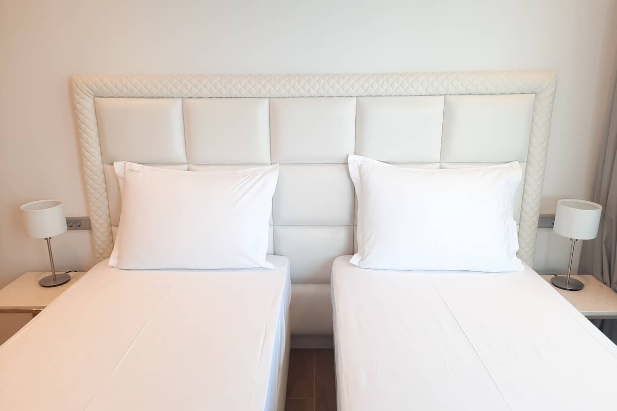 A hotel room with two single beds, each with a white mattress and pillow, and a quilted headboard. There are two bedside tables with lamps on either side of the bed.