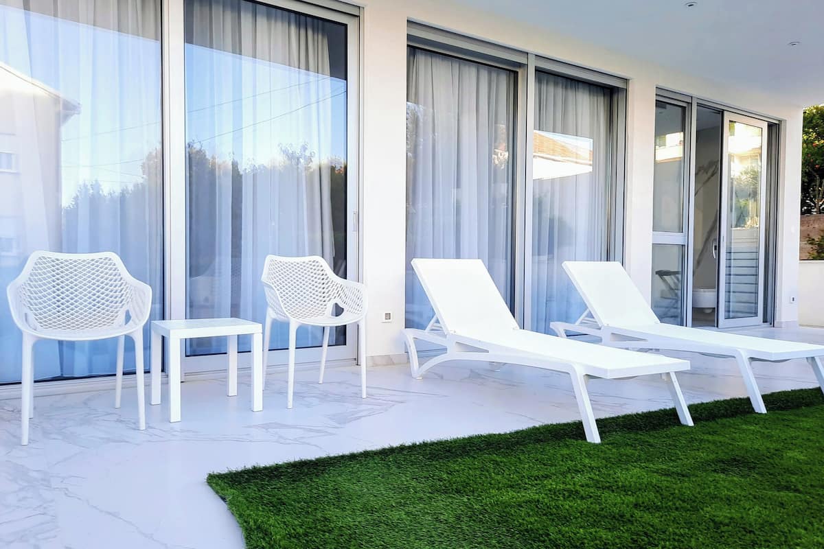 A modern outdoor seating area with white chairs and lounge chairs, large sliding glass doors, and a well-maintained lawn.