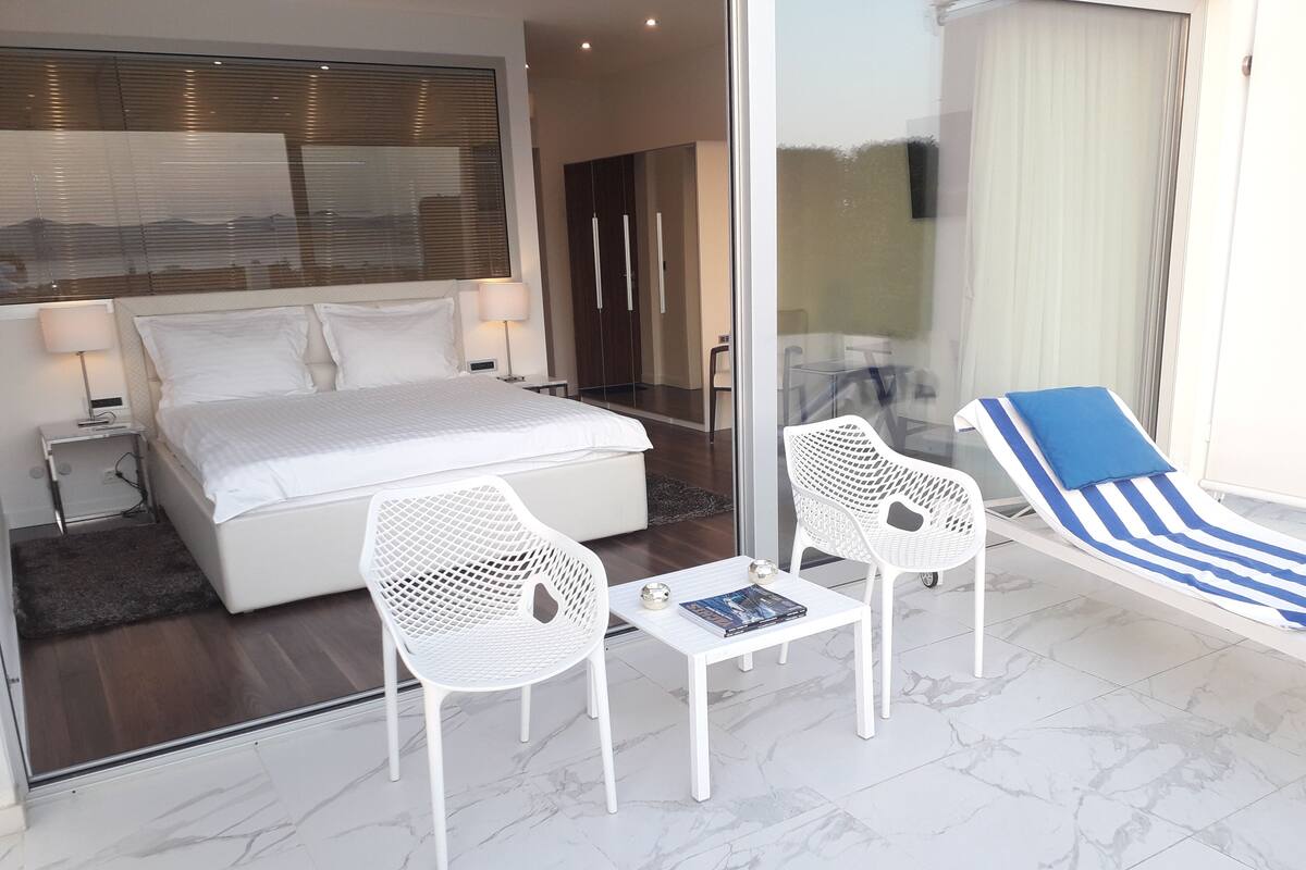 A modern bedroom with a large bed, two white chairs, a small table with a book, and a striped lounge chair.