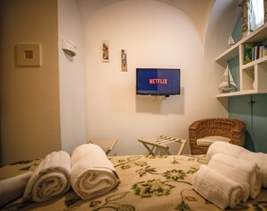 Une chambre d’hôtel avec un lit, un téléviseur affichant le logo « NETFLIX », une chaise en osier, une petite table et une étagère avec des livres et des objets décoratifs.