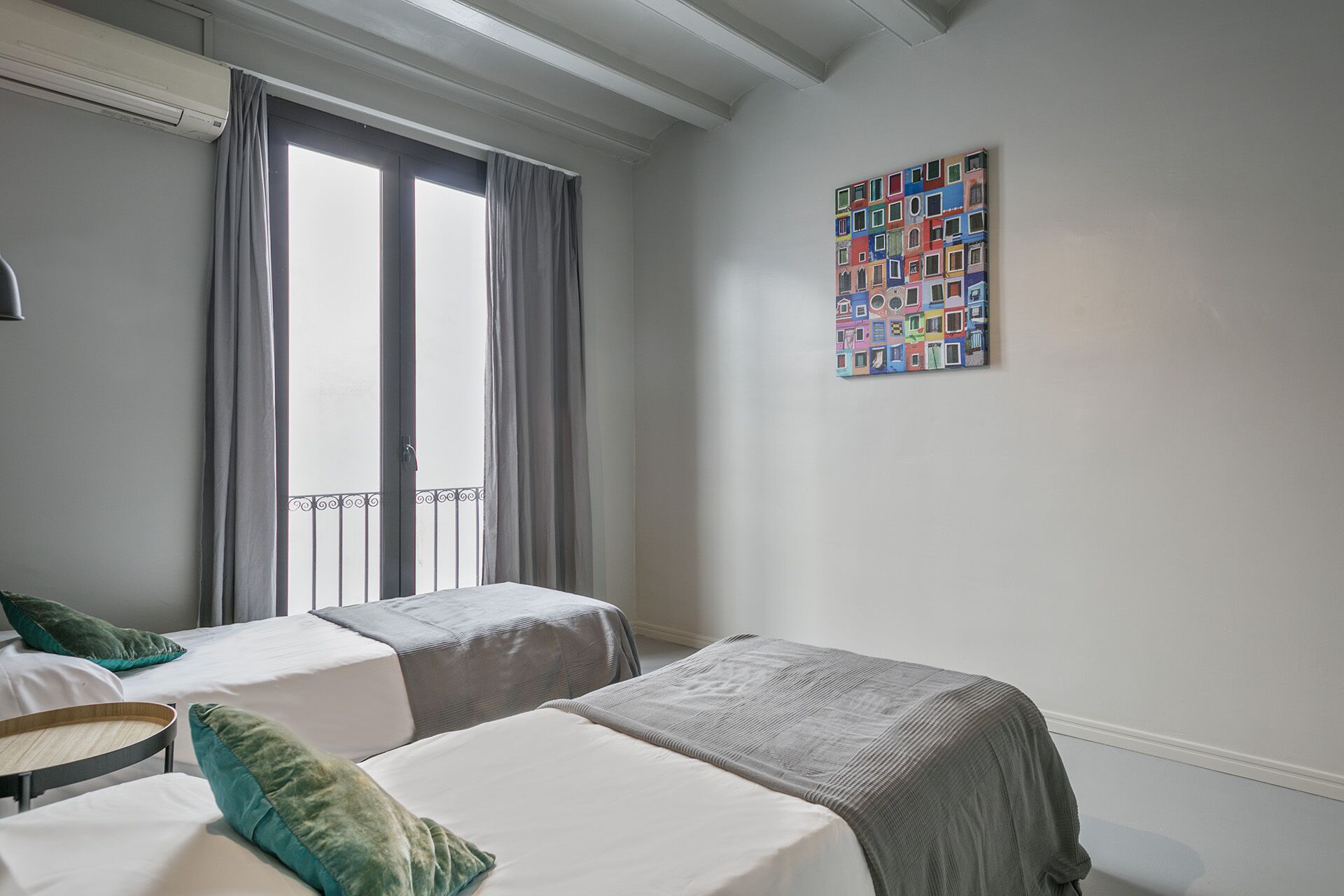 A hotel room with two single beds, a window with curtains, a balcony, and a colorful artwork on the wall.