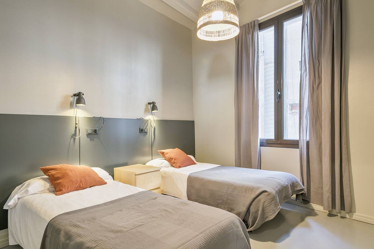 Une chambre d’hôtel avec deux lits, une grande fenêtre avec des rideaux et des lampes fixées au mur.