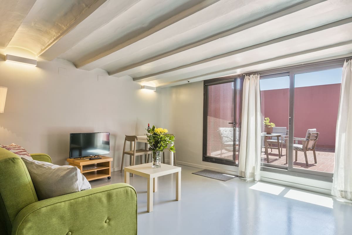Un salon moderne avec un canapé vert, un téléviseur, une petite table sur laquelle est posé un vase, et une porte-fenêtre coulissante donnant sur une terrasse extérieure.