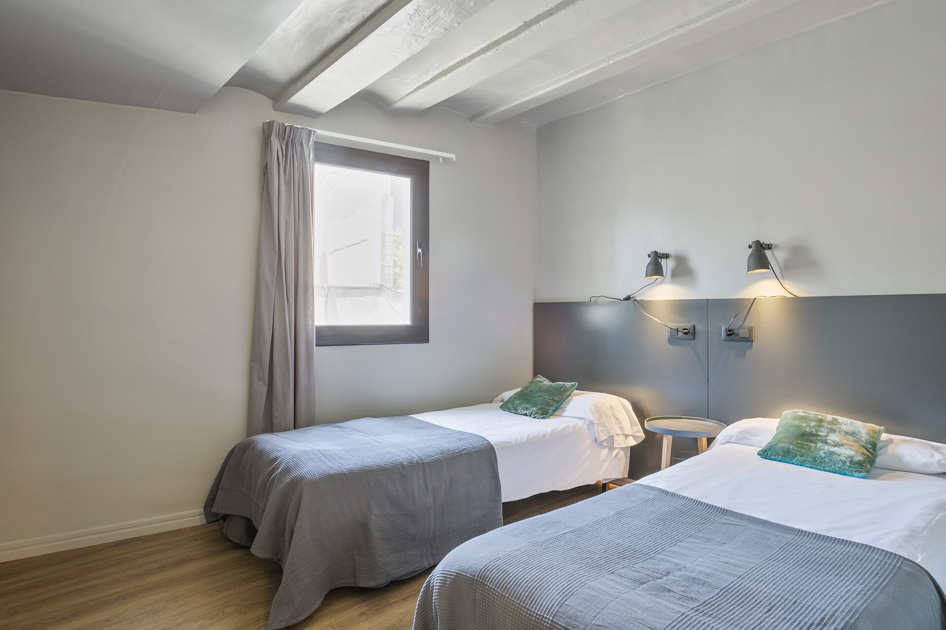 A bedroom with two beds, a window with curtains, and wall-mounted reading lamps.