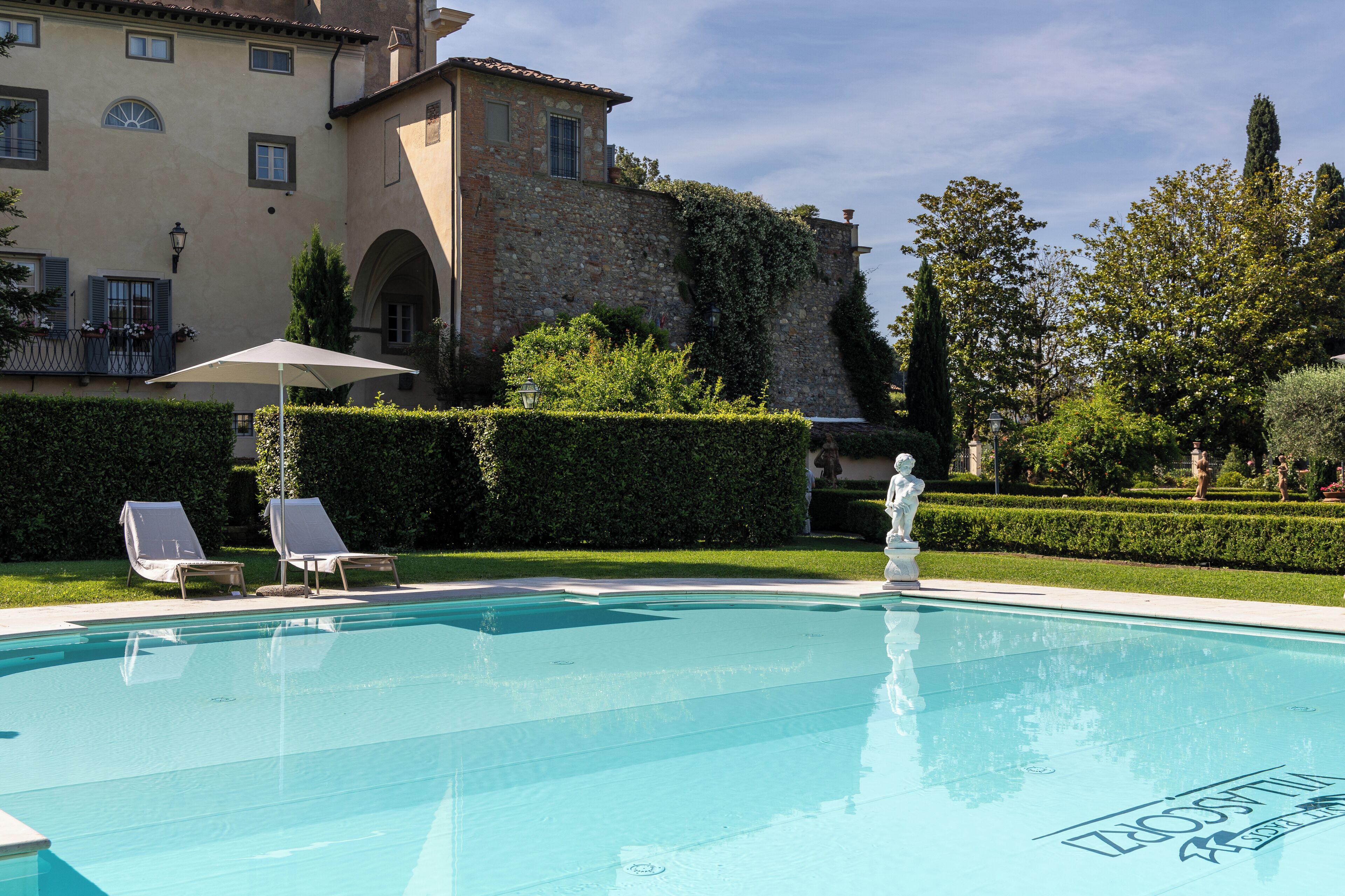 Una piscina con lettini e una statua, circondata da siepi curate e un grande edificio con pareti in pietra.