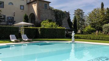Una piscina con lettini e una statua, circondata da siepi curate e un grande edificio con pareti in pietra.