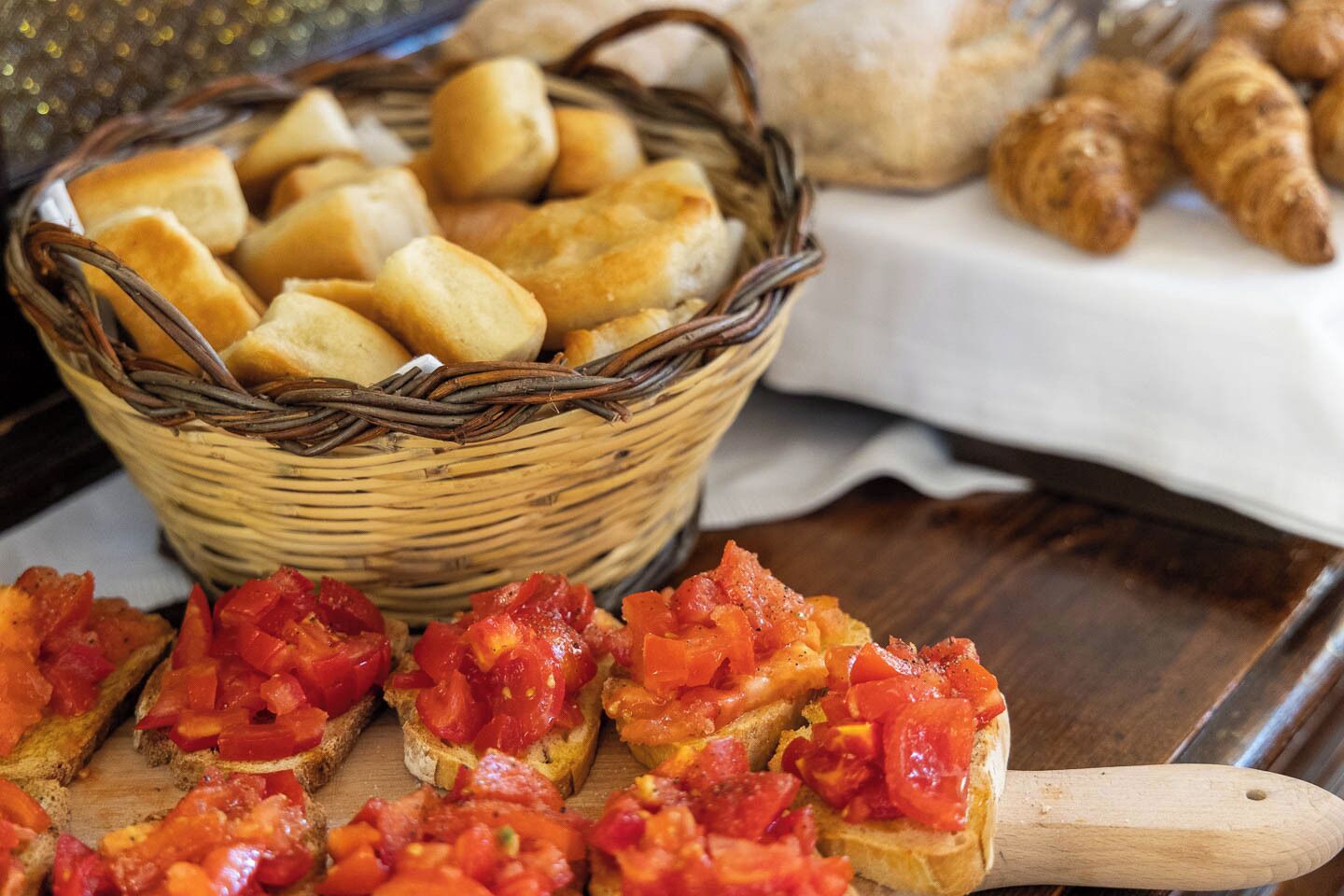 A basket of bread rolls, a wooden cutting board with tomato slices, and a wooden-handled knife.