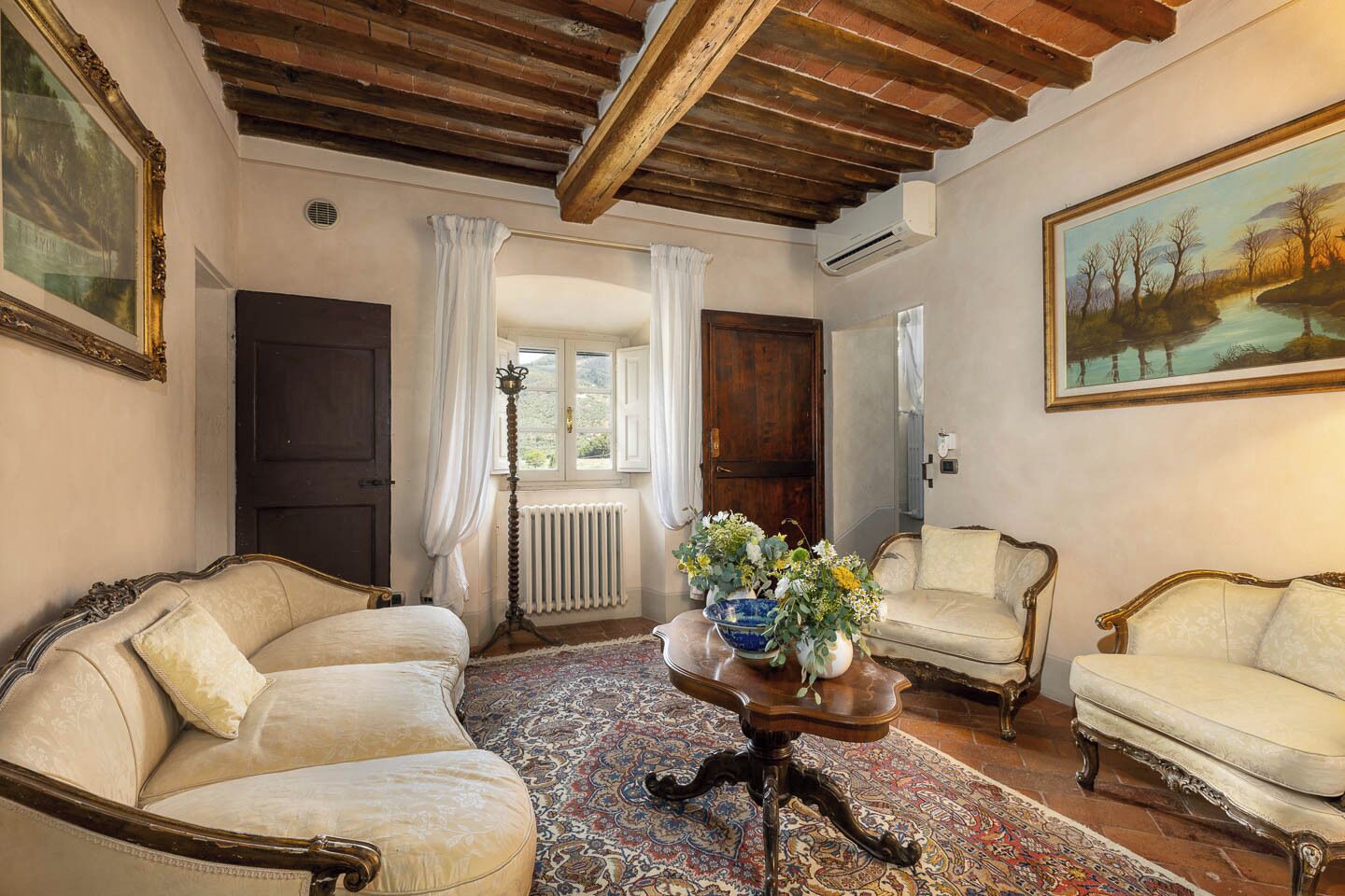 Ein Zimmer im traditionellen Stil, mit Holzbalkendecke, Heizkörper, großem Fenster mit Vorhängen und einem Gemälde an der Wand.