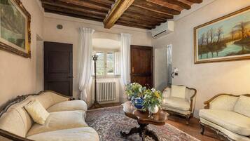 Ein Zimmer im traditionellen Stil, mit Holzbalkendecke, Heizkörper, großem Fenster mit Vorhängen und einem Gemälde an der Wand.