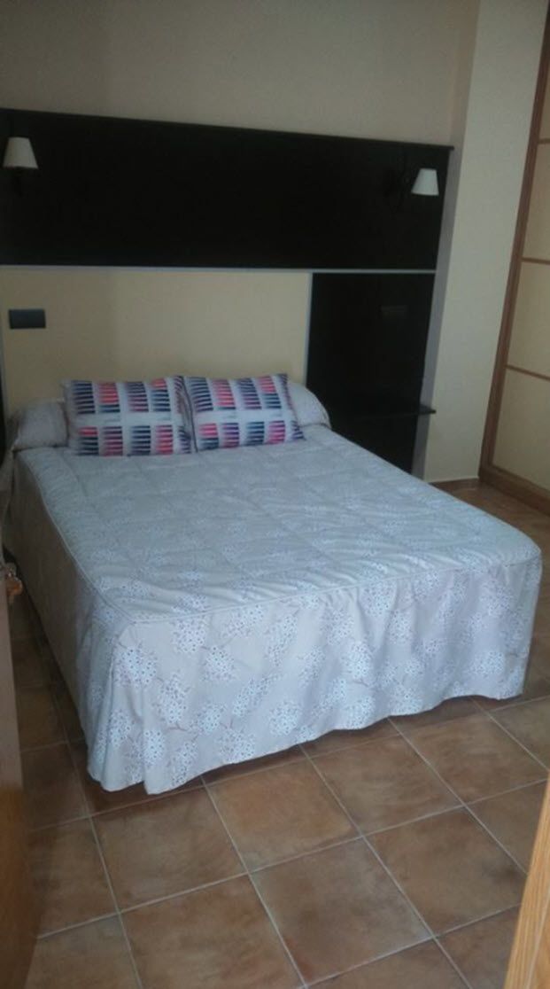 Una cama con cubrecolcha blanco, almohadas estampadas, cabecera negra y piso de baldosas.