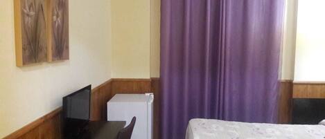 Quarto de hotel com cama, escrivaninha com computador, pequena geladeira e janela com cortinas roxas.