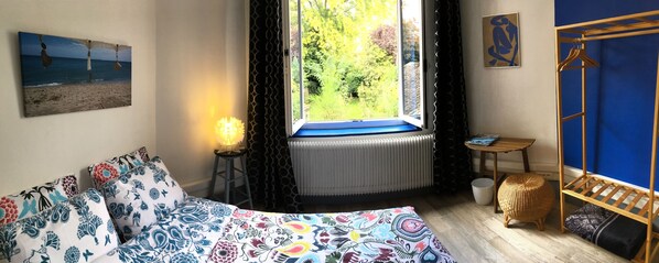 A bedroom with a bed, a window with a view of greenery, a radiator, and a wooden shelf. - La Maison du Bulot (Fruges)