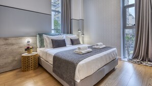 Una habitación de hotel moderna con una cama grande, una mesita de noche con un florero y vistas a la ciudad a través de amplios ventanales.