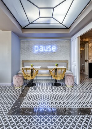 A modern interior with a geometric ceiling, a neon 'pause' sign, two wicker chairs, a cushioned bench, and patterned flooring.