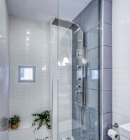 Egy modern fürőszoba üvegfalas zuhanyzóval, fehér csempézett falakkal, egy kis ablakkal és egy WC-papírtartóval.