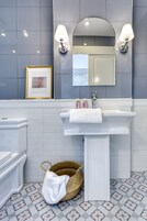 A bathroom with a white sink, two wall-mounted lamps, a framed picture above the mirror, and a woven basket with a white towel on the floor.