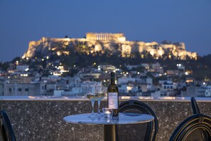 Una mesa con una botella de vino y copas, con vistas a un paisaje urbano y una estructura destacada en la cima de una colina.