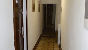 Un couloir étroit, dont le sol est recouvert de carreaux décoratifs, avec une porte en bois sur la gauche et un luminaire fixé au mur.