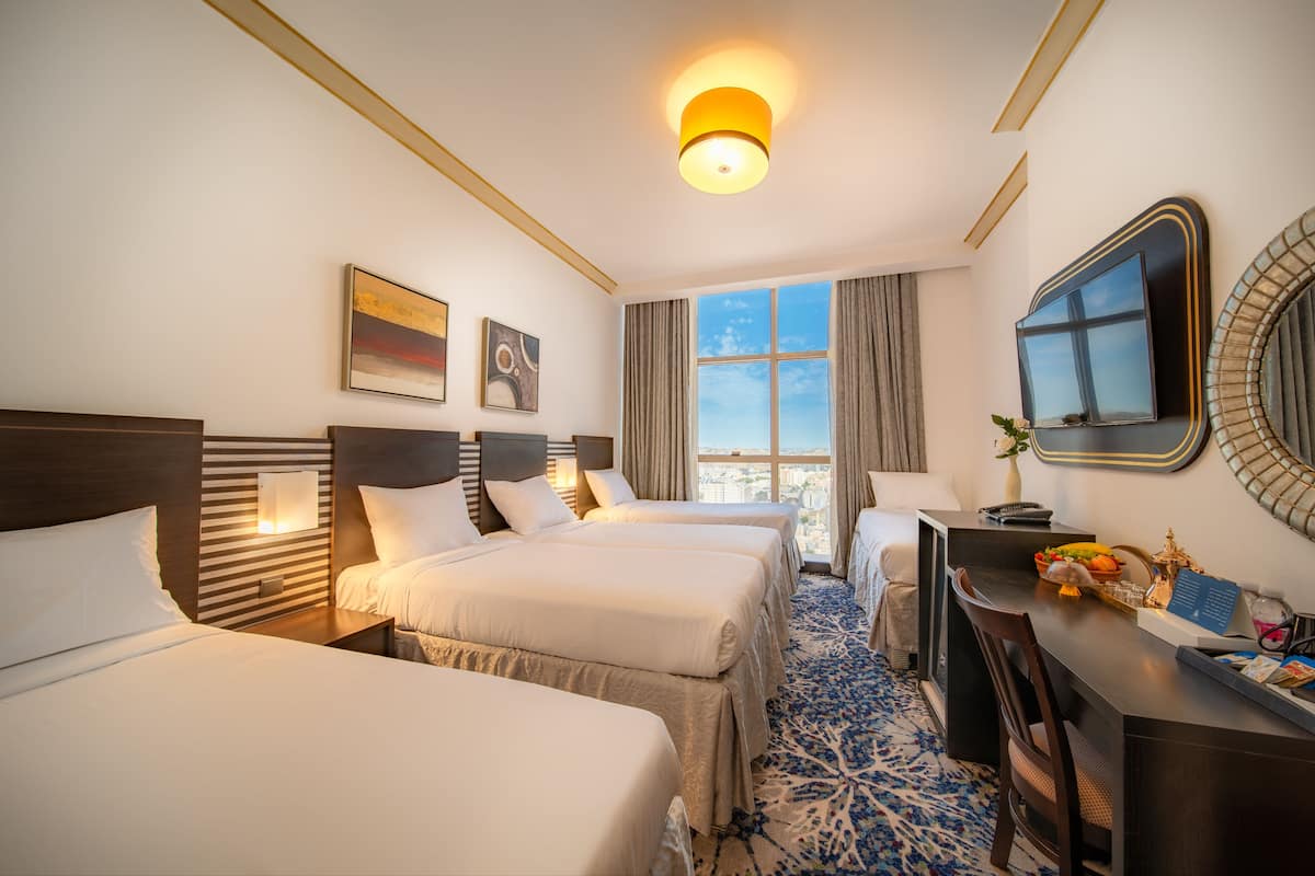 A hotel room with three beds, a desk, a chair, a large window with curtains, and a city view.