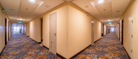 A long, narrow hotel corridor with patterned carpet, doors on both sides, and overhead lighting.