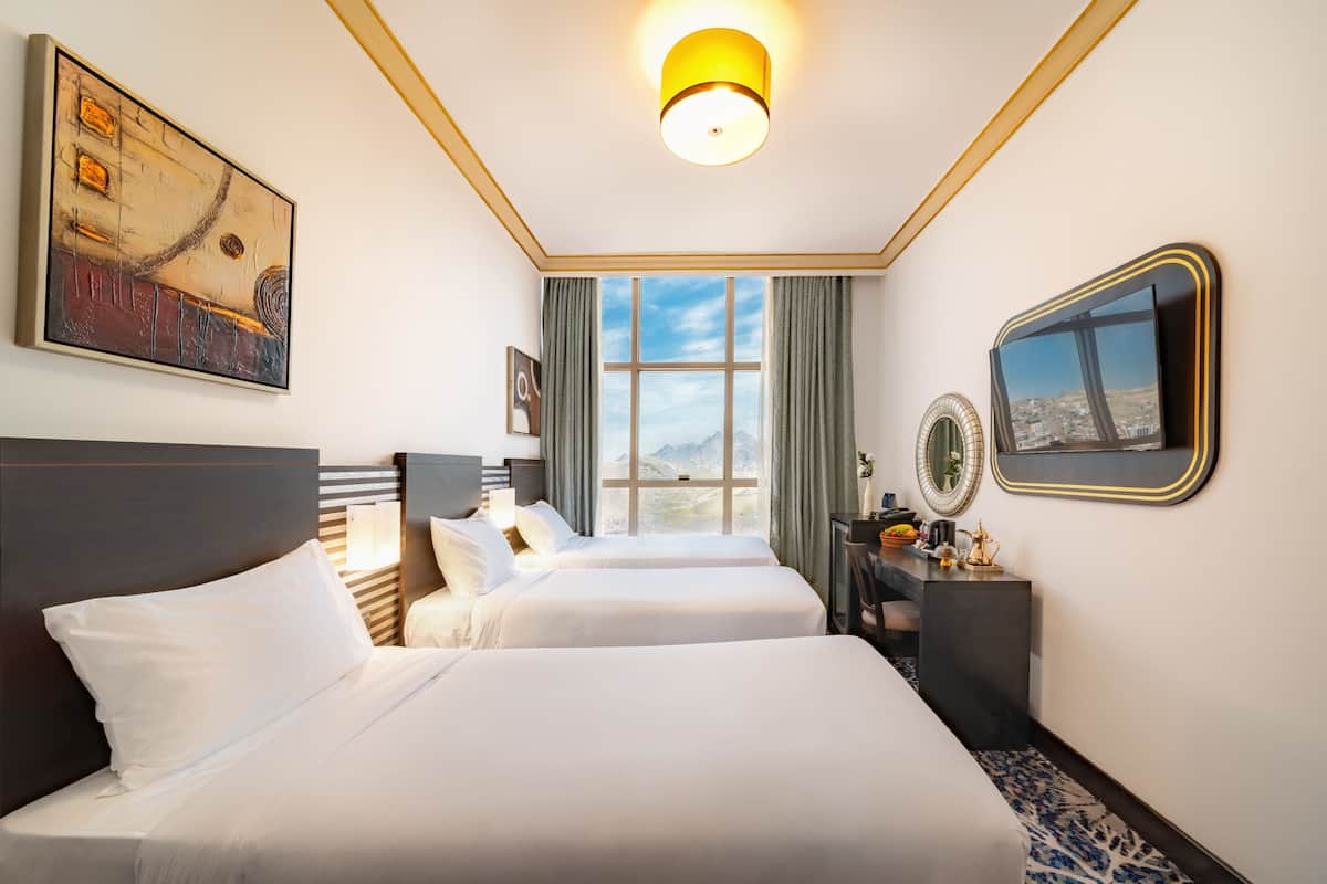 A hotel room with two beds, a large window with curtains, a painting, a mirror, and a desk with a lamp.