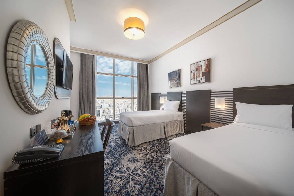 A hotel room with a bed, a desk with a telephone, a decorative bowl, and a large window offering a city view.