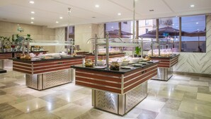 Una cocina moderna de hotel con múltiples estaciones de buffet, pisos de mármol y ventanas amplias que ofrecen vistas al exterior.
