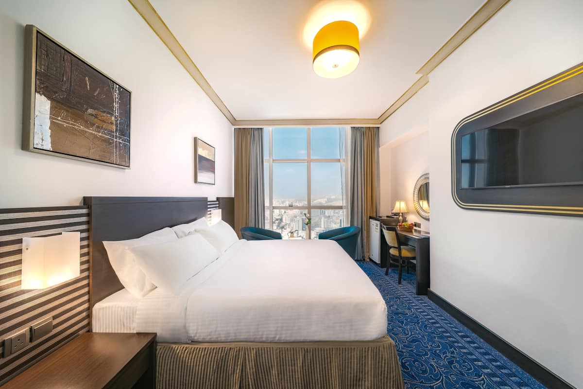A hotel room with a large bed, a desk, a chair, a window with a city view, and a painting on the wall.