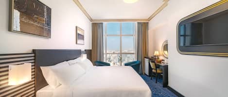 A hotel room with a large bed, a desk, a chair, a window with a city view, and a painting on the wall.
