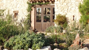 A stone building with a wooden door, surrounded by various plants and rocks. - Mas de l'Arlequi Casa rural (Montblanc)