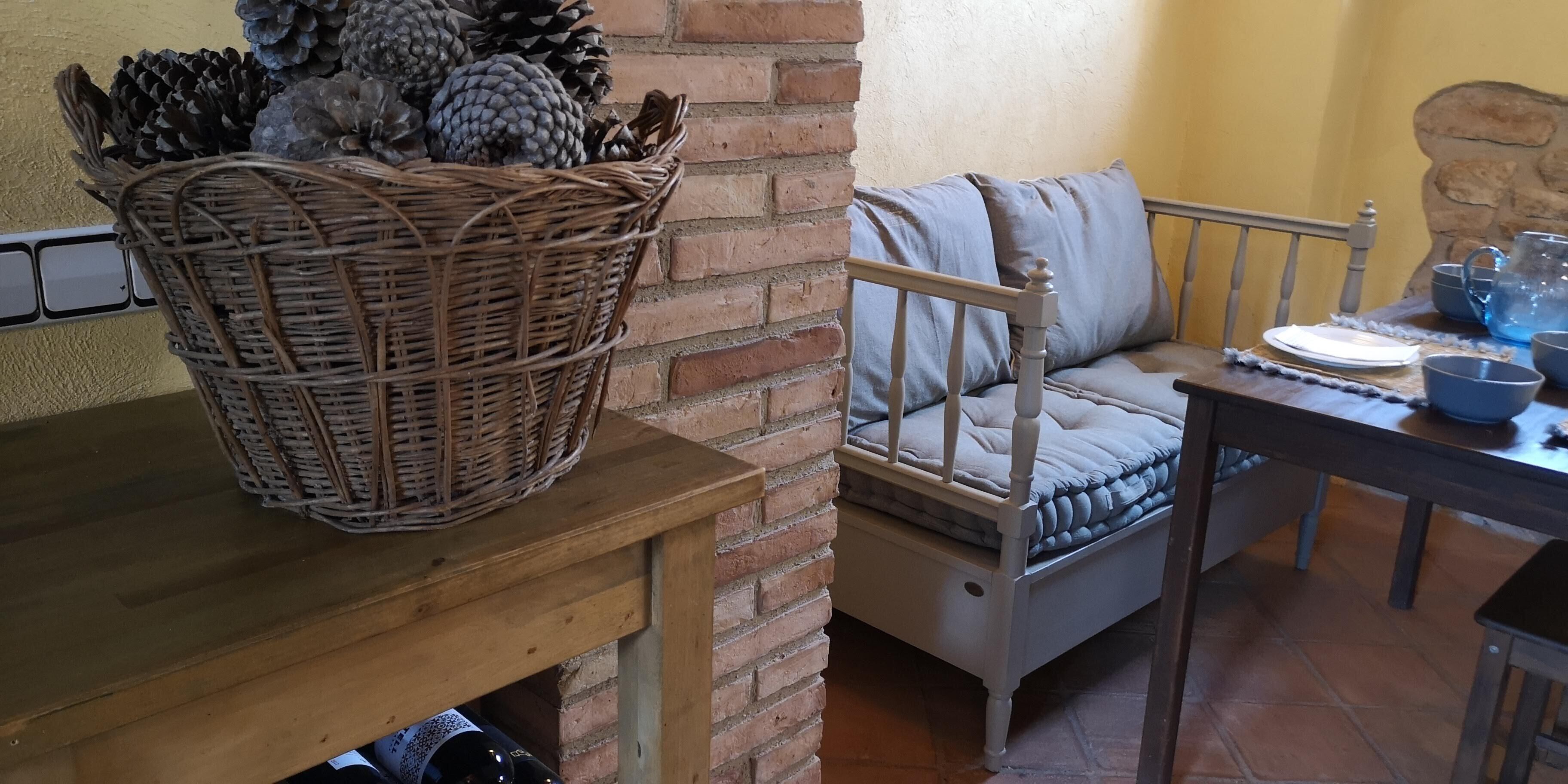 Una habitación acogedora con una cesta de mimbre llena de piñas sobre una mesa de madera, un banco con cojín y una mesa de comedor dispuesta con platos, tazas y una jarra.