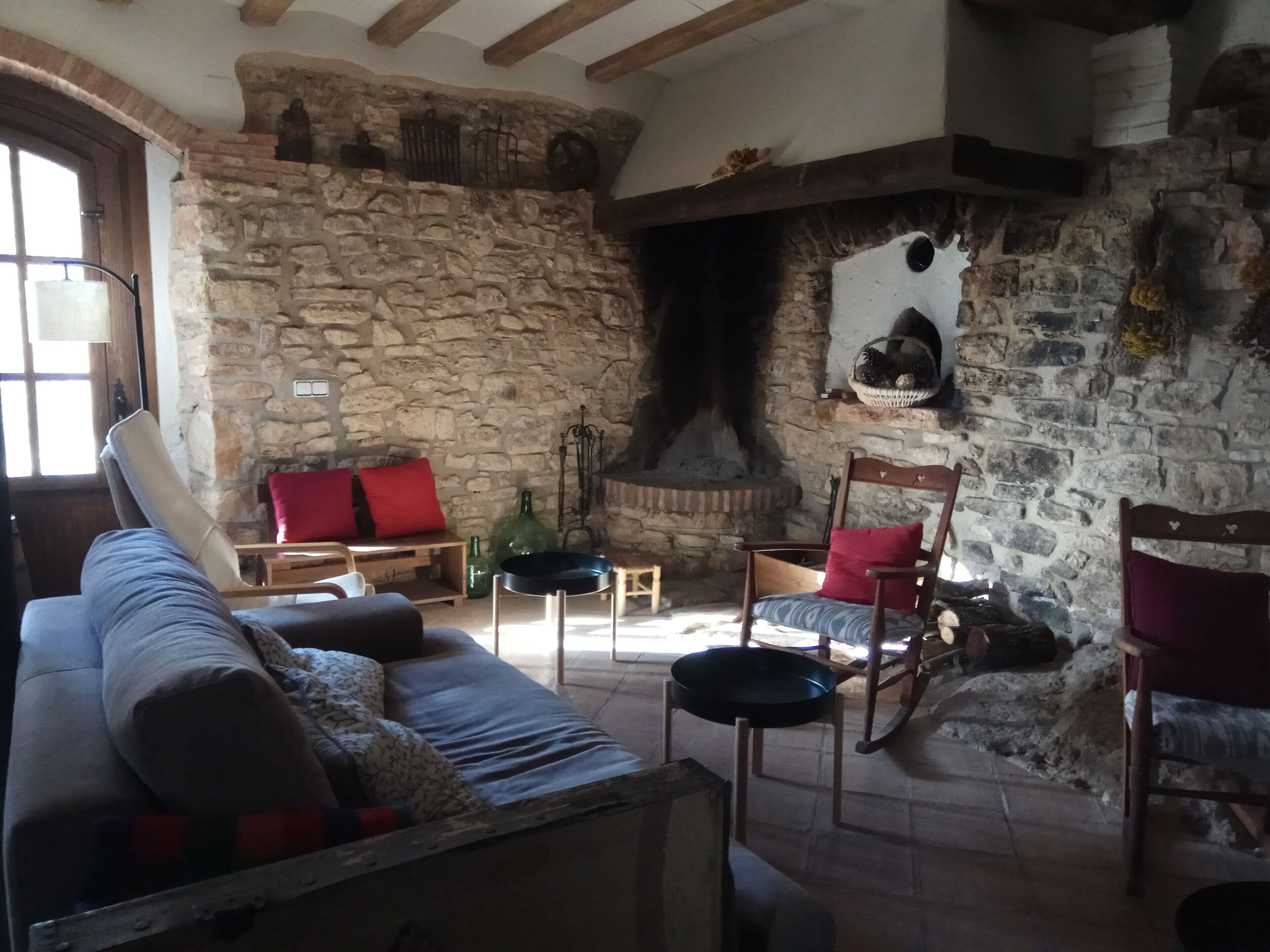 Um quarto rústico com paredes de pedra, lareira, um banco de madeira, um sofá com almofadas vermelhas e uma mesa de jantar com cadeiras.