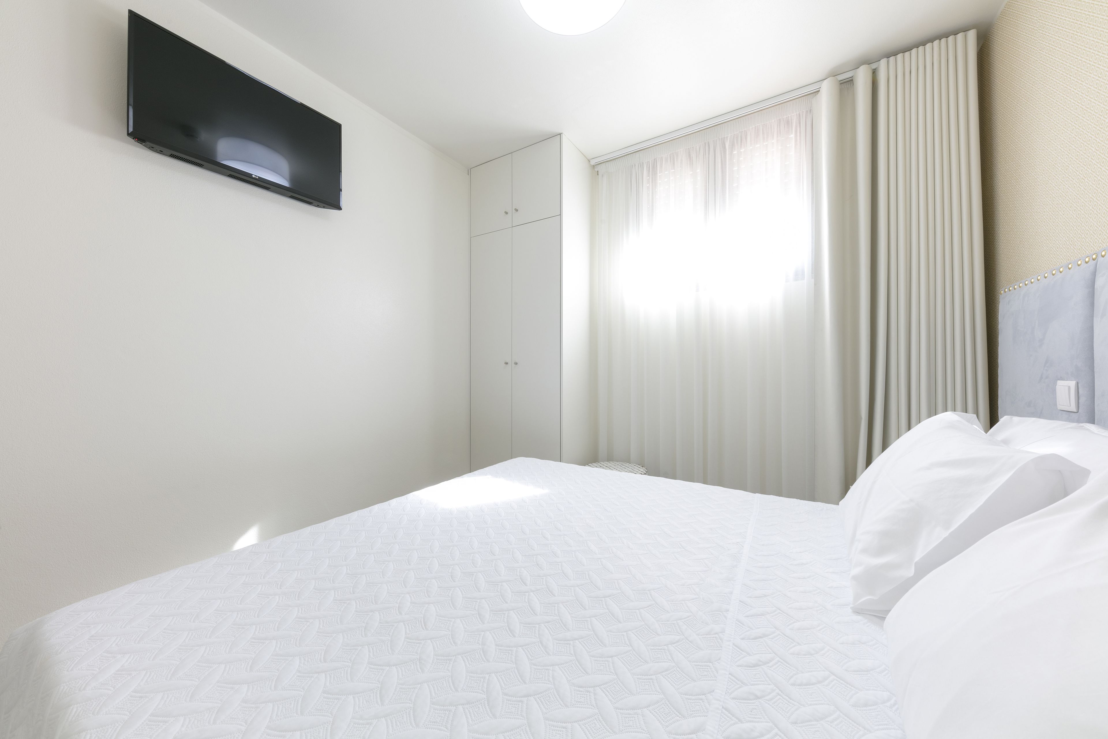A hotel room with a bed, a flat-screen TV mounted on the wall, and a window with curtains.
