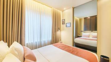 Quarto de hotel com uma cama grande, cortinas bege, uma mesa de cabeceira com abajur e um espelho refletindo a cama.