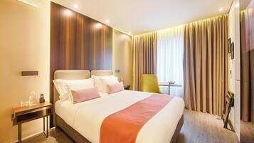 Habitación de hotel con una cama grande, una mesita de noche con lámpara, una silla y vistas al exterior a través de cortinas.