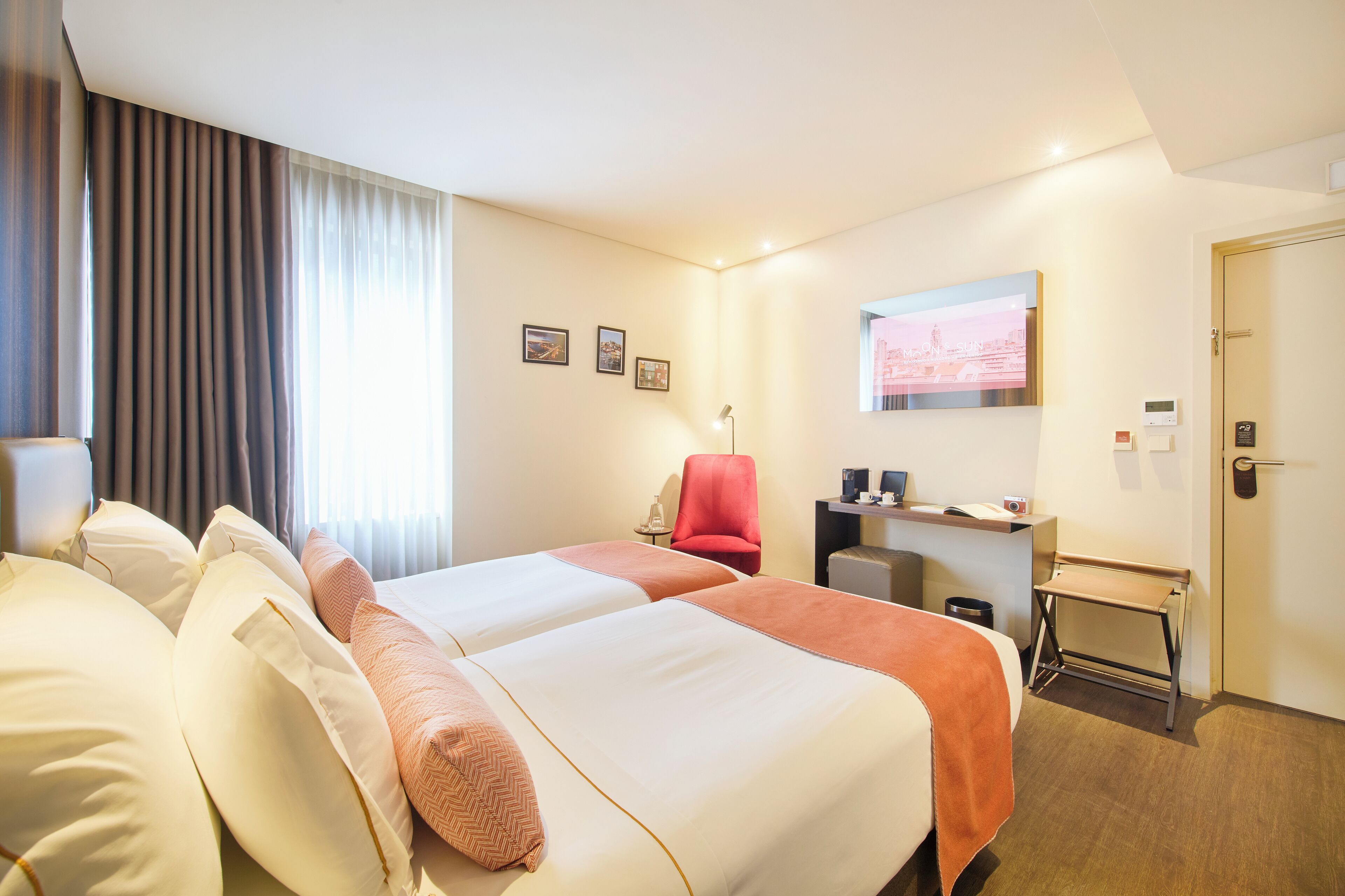 A hotel room with two beds, a desk, a chair, a TV, and a door.