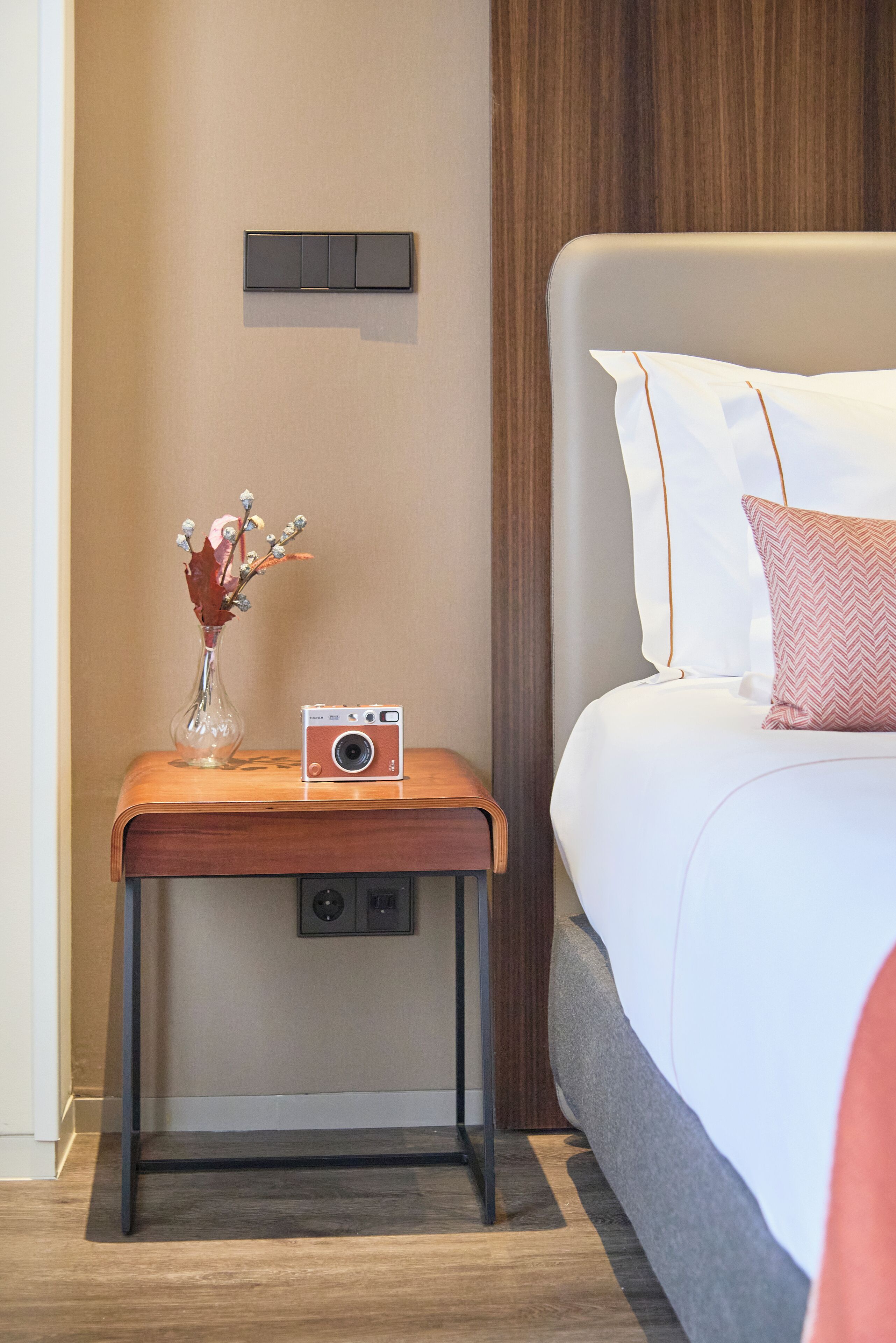 A neatly arranged hotel room with a bed, a bedside table with a camera, and a vase with dried flowers.