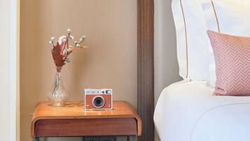 A neatly arranged hotel room with a bed, a bedside table with a camera, and a vase with dried flowers.